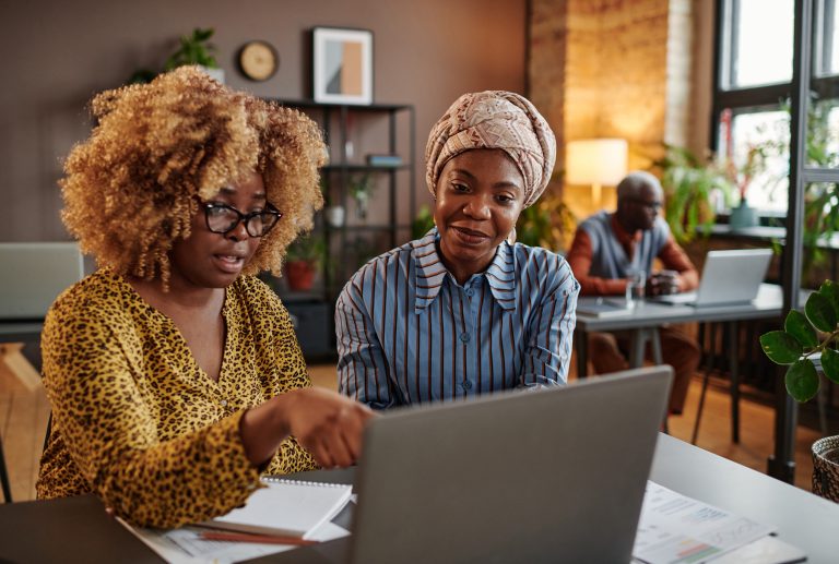 African businesswoman with curly hair pointing at laptop while sitting at the table with her colleague and they working in team at office